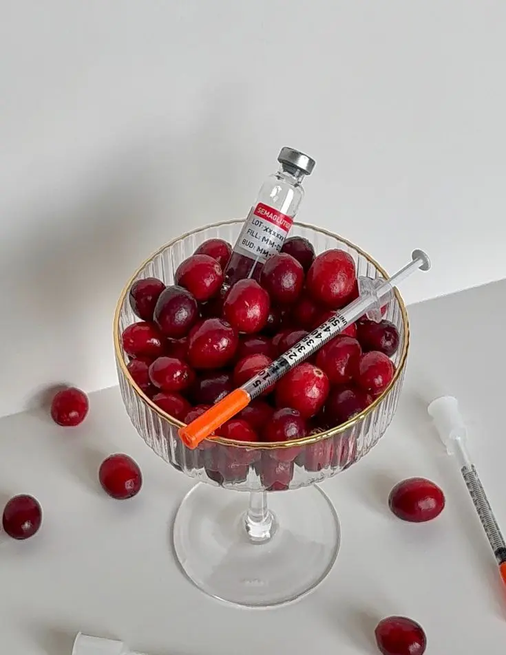 Cherries in a glass bowl with a syringe and small vial as medical-themed props in a still-life setup.